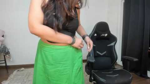 Media: Video of a woman in a black crop top and vibrant green skirt, adjusting a gaming chair, in a minimalist, carpeted room with a white wall and black curtains.