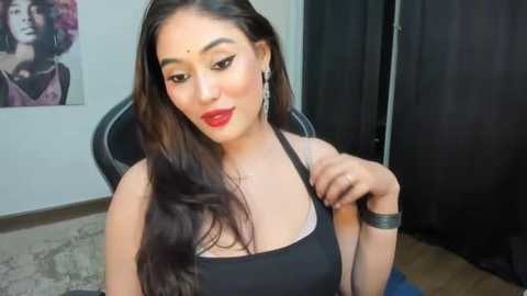 Media: Video of a young woman with long dark hair, fair skin, and red lipstick, wearing a black tank top and silver earrings, sitting indoors with a black chair and a black curtain in the background.