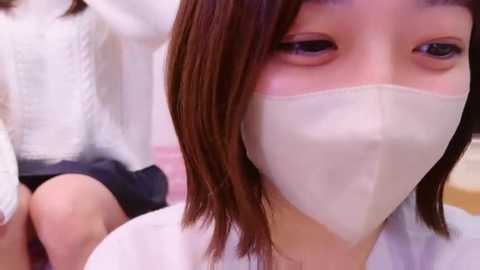 Media: Video of an Asian woman with straight, shoulder-length brown hair, wearing a white surgical mask, seated indoors. Another person, partially visible, is in the background.