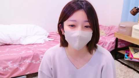 Media: Video of a young East Asian woman with shoulder-length brown hair, wearing a white face mask, a gray V-neck shirt, and sitting in a pink-patterned bedroom with a messy bed, nightstand, and blue curtains.