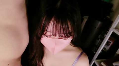 Media: A close-up video of a young Asian woman with long black hair, wearing a pink face mask and a light purple spaghetti strap top, in a dimly lit room.