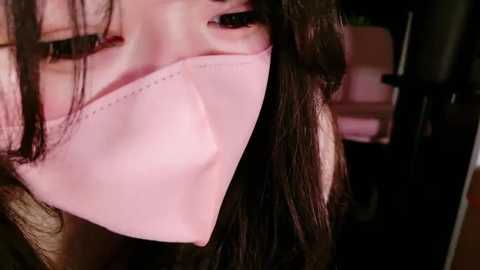 Media: Close-up video of a woman with long, dark hair partially covering her face, wearing a pink surgical mask. The background is dimly lit, showing a blurred, indoor setting.