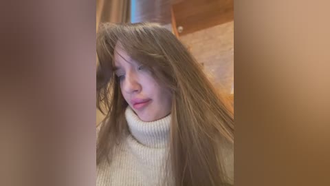 Media: Video of a young woman with long, messy brown hair, wearing a cream-colored turtleneck sweater, indoors with blurred background.