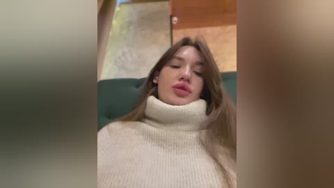 Media: Video of a young woman with light skin, long brown hair, and full lips, wearing a cream turtleneck sweater, lying on a green couch. The background shows wooden flooring and a doorway.