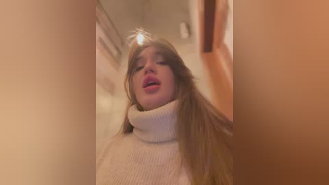 Media: A video of a Caucasian woman with long, straight brown hair, wearing a cream-colored turtleneck sweater, looking upwards in a dimly lit hallway, with a blurred background of orange and beige walls.