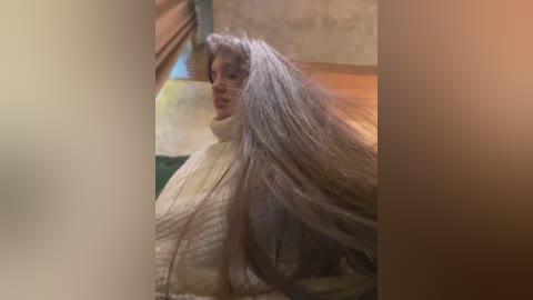 Media: Video of a woman with long, silver hair, wearing a cream-colored sweater, indoors, with a blurred background featuring a wooden door and wall.