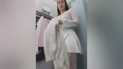 Media: Video of a young Asian woman in a white lace dress, holding a white blanket, standing in a changing room with pink and black garments hanging nearby.