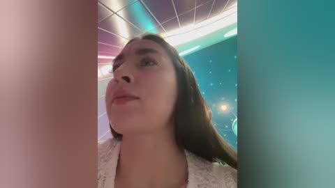 Video of a young woman with light brown skin and long brown hair, wearing a white lace top, standing in a futuristic room with blue and pink lights and a tiled ceiling.