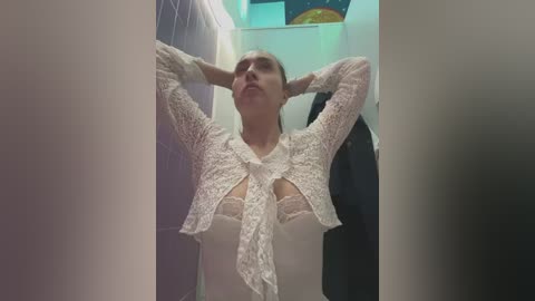 Video of a woman in a white lace top with cutout details, hands behind head, standing in a dimly lit, cluttered closet with hanging clothes.