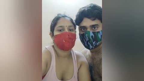 Media: Video of a South Asian couple in a room, both wearing red face masks and casual clothing; the man has a camouflage-patterned mask and the woman a sleeveless pink top.