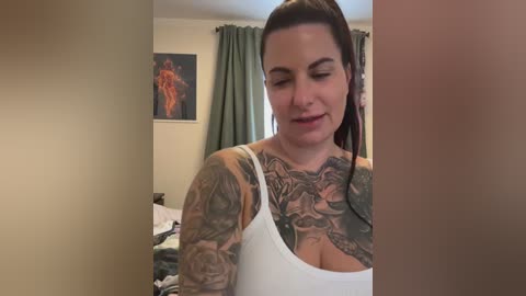 Media: Video of a tattooed woman with fair skin, long dark hair tied back, wearing a white tank top, standing in a bedroom with green curtains and a framed painting on the wall.