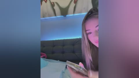 Media: Video of a young woman with long, straight, light brown hair, reading a book in a dimly lit bedroom with a green and blue glow, a dark green headboard, and a red item on the bed.