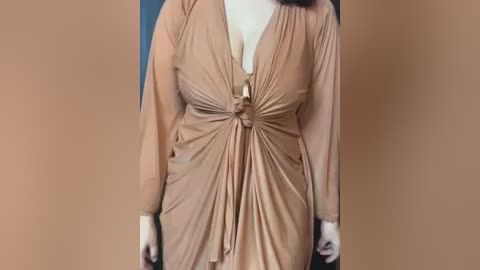 Media: Video of a woman wearing a flowing, light brown, V-neck dress with a gathered waist, revealing a white shirt underneath. The background is a solid, light brown.