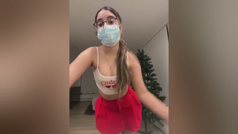 Media: Video of a young woman with fair skin, wearing a light blue face mask, glasses, a \"Chili's\" tank top, and a red skirt, standing in a modern, minimalist room with a Christmas tree in the background.