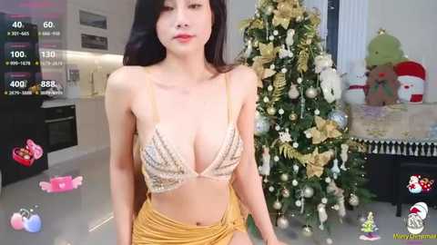 Media: Video of an East Asian woman with long black hair, fair skin, and a slim figure, wearing a glittery bra and gold skirt, posing in a modern, festive living room with a decorated Christmas tree.