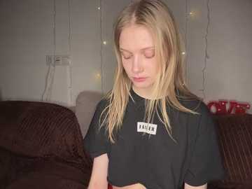 Media: A video of a young Caucasian woman with long, straight blonde hair, wearing a black t-shirt with a white name tag, seated on a dark brown couch in a dimly lit room with string lights and a red Christmas ornament in the background.