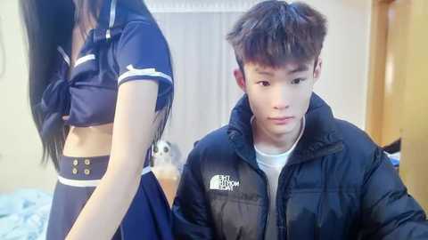 Media: Video of a young East Asian man in a puffer jacket, holding a drink, with a woman in a revealing sailor outfit and a stuffed toy in the background.