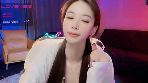 Media: A video of a fair-skinned, long-haired Asian woman with closed eyes, wearing a white top, in a dimly lit room with a purple and blue light effect, lounging on a black sofa.