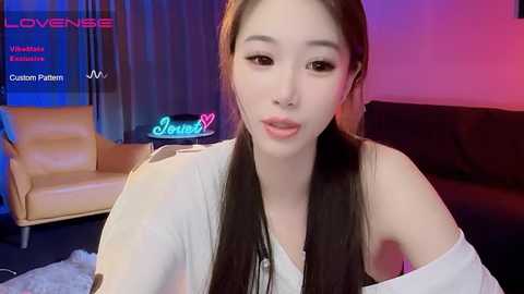 Media: Video of an East Asian woman with long, straight black hair and fair skin, wearing a white off-shoulder top, sitting indoors with a light brown leather armchair and colorful neon \"Love\" sign in the background.