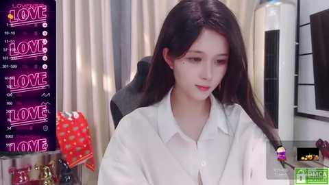 Media: Video of a young East Asian woman with long black hair, fair skin, and light makeup, wearing a white button-up shirt. Background includes a screen with \"LOVE\" text, a red polka-dotted towel, and a modern living room with beige curtains and black furniture.