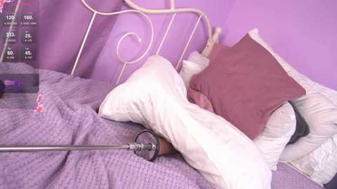 Media: Video of a purple-themed bedroom with a white wrought-iron bed, white pillow, and a brown pillow, covered in a lavender quilt. A metal vibrator is partially visible on the bed, suggesting a sexual context.