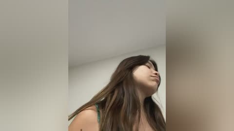 Media: Video of a young woman with long, dark brown hair, wearing a green tank top, standing in a dimly lit room with white walls. Her head is tilted back, eyes closed, and she appears to be in deep thought.