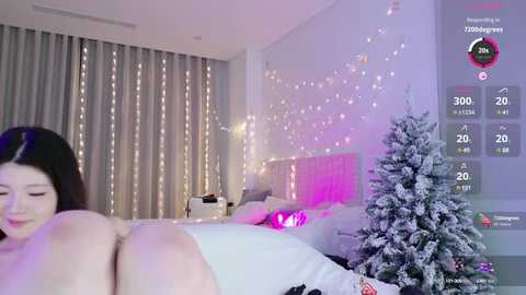 Media: A video of an Asian woman with long black hair, lying on a bed, wearing a white shirt, in a softly lit bedroom with a Christmas tree and string lights.