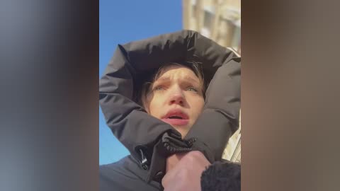 A video captures a young, light-skinned boy with short brown hair, wearing a black winter coat, reaching up, his mouth slightly open. The background features a clear blue sky and a beige building. The boy's expression is one of surprise or excitement.