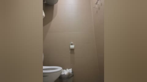 Media: A video of a compact, beige-tiled bathroom with a white toilet and a roll of toilet paper on the wall, positioned in the foreground. The room is dimly lit, creating a subdued atmosphere.