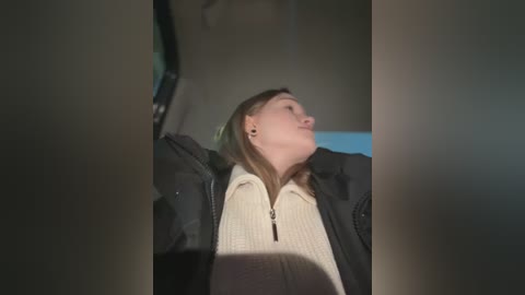 Media: A video of a woman with light skin and long brown hair, wearing a cream sweater under a black jacket, seen from a low angle, leaning back in a car.