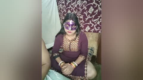 Media: A video of a woman in a maroon sari with intricate embroidery, seated on a bed, wearing a purple mask and multicolored bangles. The background features patterned maroon and white wallpaper.