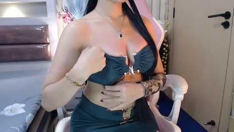 Media: A video of a woman with long black hair, fair skin, and a slender physique, wearing a dark green strapless bra and high-waisted pants, sitting on a white chair in a modern, minimally decorated room.