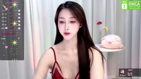 Media: Video of a slender East Asian woman with long black hair, fair skin, and red lipstick, wearing a red lace camisole, against a soft white backdrop with a pink piggy bank and flowers.