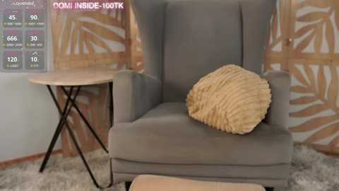 Media: Video of a modern, gray upholstered armchair with a textured, beige throw pillow, set against a wall with a tropical leaf-patterned wallpaper. A small, round wooden side table with black legs stands nearby.