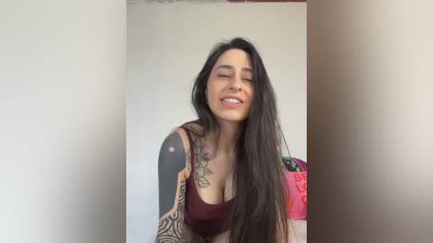 Media: Video of a smiling Latina woman with long black hair, wearing a burgundy tank top, showcasing tattoos on her left arm and chest, against a plain beige wall.