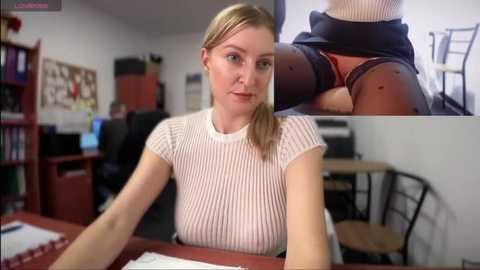 Media: Video of a blonde woman with fair skin and a slender build, wearing a white ribbed top, seated at a desk in an office. A close-up inset shows her in black lingerie.