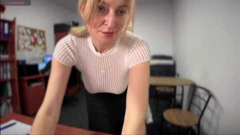 Media: Video of a Caucasian woman with light skin, blonde hair, and fair complexion, wearing a white ribbed t-shirt and black skirt, leaning over a desk in a cluttered office with bookshelves and a computer monitor.