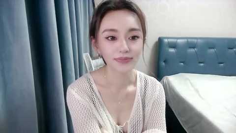 Media: Video of an East Asian woman with fair skin and straight black hair, wearing a white, knitted cardigan, sitting on a bed with a blue headboard and white sheets, in a modern, minimalistic bedroom.