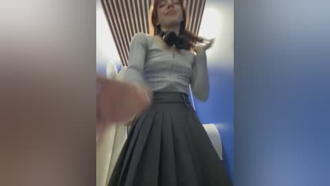 Media: A low-angle video of a young woman in a school uniform, wearing a white blouse and a grey pleated skirt, with a striped wall and a blurred background.