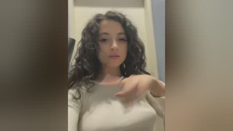 A video of a young woman with curly dark hair, fair skin, and medium breasts, wearing a beige long-sleeved shirt, in a dimly lit room, making a \"thumbs-up\" gesture.