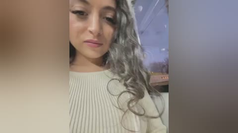 Video of a young woman with medium skin tone, curly gray hair, wearing a cream ribbed sweater. Background shows a blurred indoor setting with soft lighting.