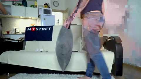 Media: Video of a person in a grey shirt and blue jeans, holding a large, gray paddle, standing in a modern kitchen with white cabinets, a microwave, and a white \"MST BET\" banner on a sofa.