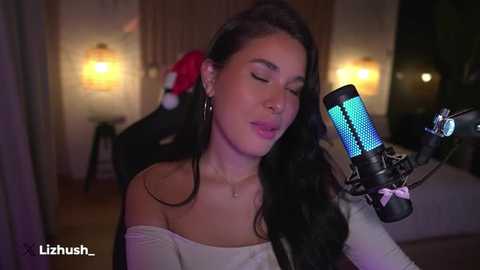 Media: Video of a young woman with long black hair, wearing a white off-shoulder top, singing into a blue LED microphone in a dimly lit room with warm lighting and a bed in the background.