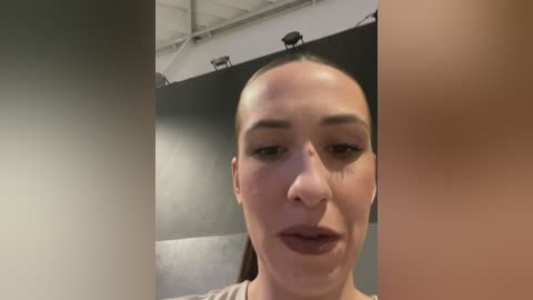 Media: Video of a bald, light-skinned woman with minimal makeup, wearing a light-colored top, standing in an indoor studio with a grey wall and ceiling lights.