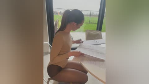 Media: A video of a young woman with light skin, dark hair in a ponytail, wearing a beige sweater and black shorts, sitting on the floor studying a large sheet of paper on a wooden table, inside a modern, brightly lit room with large windows.