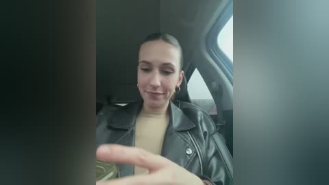 Media: Video of a young woman with light skin and straight dark hair, wearing a black leather jacket over a beige top, smiling and holding a phone, seated in a car with grey seats.