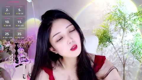 Media: Video of an Asian woman with long black hair, fair skin, and red lipstick, wearing a red top, sitting in a room with a green plant and a calendar displaying 25.