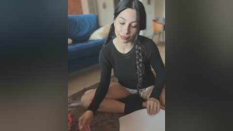 Media: A video of a woman with long black hair, wearing a black top and white shorts, sitting cross-legged on a rug, focusing on a paper.