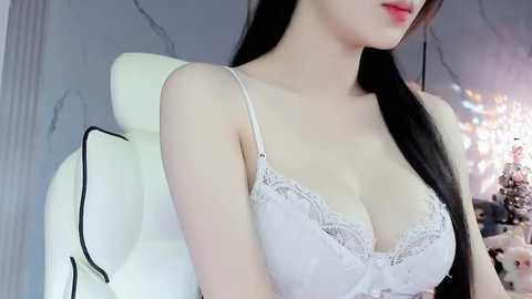 Media: Video of a fair-skinned woman with long black hair, wearing a white lace bra, sitting on a white gaming chair, in a dimly lit room with soft lighting and a Christmas tree in the background.