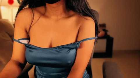 Media: Video of a woman with long, straight black hair, wearing a strapless, dark blue dress with one strap slipping off her shoulder. The background features a beige couch, a TV, and a blurred figure.